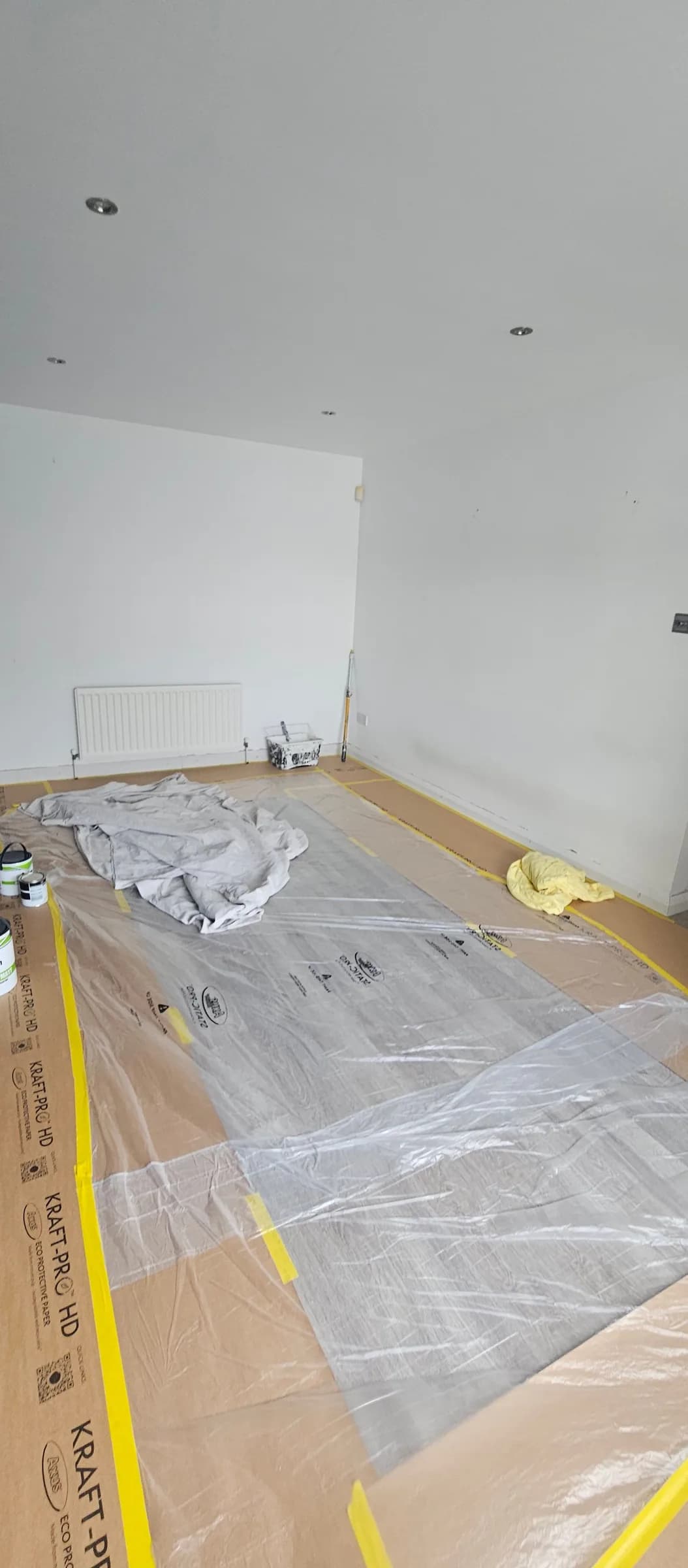 A room floor covered with Kraft-Pro paper and clear plastic sheeting, taped down along the skirting with yellow low-tack tape.