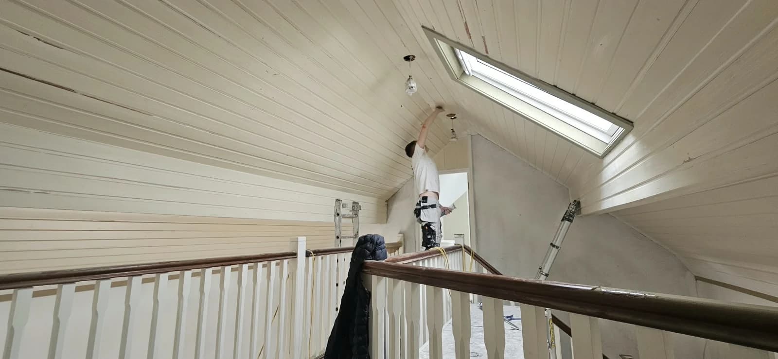 A painter reaching overhead to cut in a vaulted tongue-and-groove boarded ceiling above a galleried landing, beside a rooflight